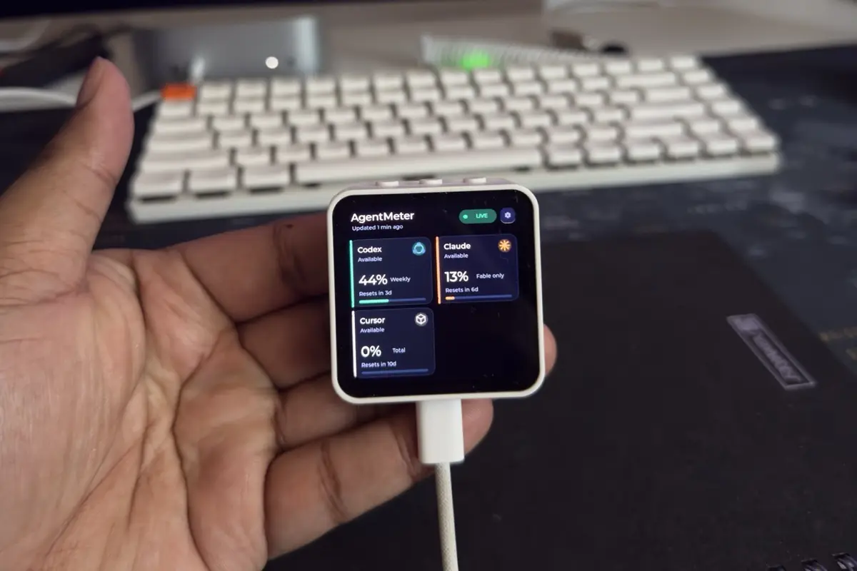 AgentMeter held in a hand above a desk, its square AMOLED screen showing usage cards